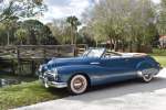 a blue classic Buick convertible parked next to a stream with a wooden bridge in the background